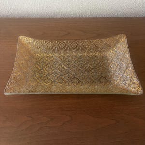 May include: A rectangular glass tray featuring a gold-toned finish and a repeating embossed quatrefoil pattern. The tray has slightly curved edges and a textured surface that creates a metallic appearance across the entire piece.