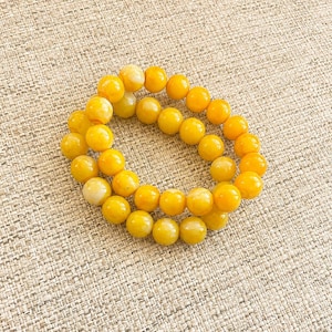 May include: A circular bracelet composed of numerous smooth, round, yellow beads. The beads exhibit varying shades of opaque and semi-translucent yellow, creating a consistent color palette throughout the piece. The beads are strung together in a continuous loop.