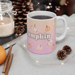 May include: A white ceramic mug with a gradient background transitioning from light orange to soft pink. The mug features a repeating pattern of watercolor-style pumpkins in shades of orange and cream, with the word Pumpkin printed in white.