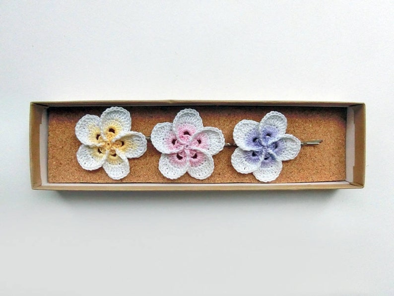 May include: Three crocheted flower hair clips in a box. The flowers are white with yellow, pink, and purple centers.