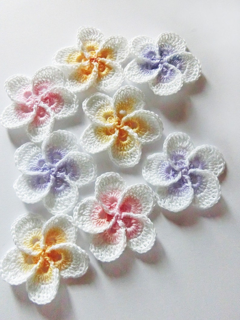 May include: Eight crocheted flowers in various pastel colors, including pink, yellow, and purple. The flowers are made with white yarn and have a layered, 3D effect.