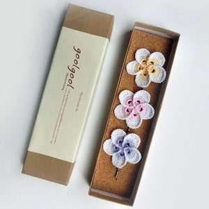 May include: Set of three crocheted flower hair clips in a brown box. The flowers are white with pink, yellow, and purple accents. The box has a label that says "goo/goo!" and "Handmade by"