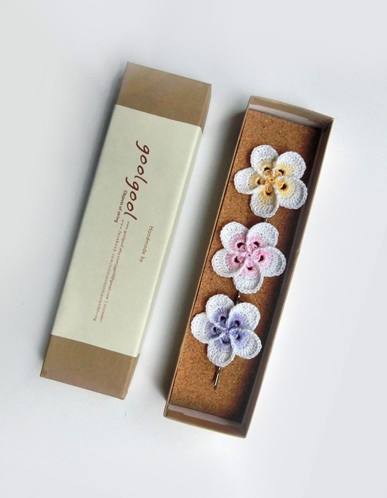 May include: Set of three crocheted flower hair clips in a brown box. The flowers are white with pink, yellow, and purple accents. The box has a label that says "goo/goo!" and "Handmade by"