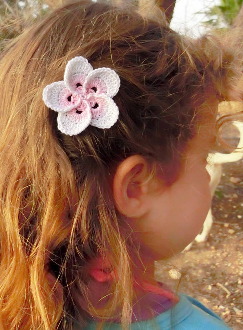 May include: A pink and white crocheted flower hair clip. The flower has five petals and is attached to a metal clip.