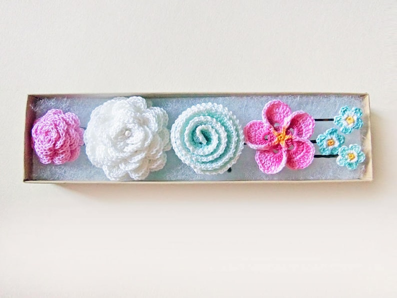 May include: A set of five crocheted flower hair clips in a box. The clips are in shades of pink, white, and blue.