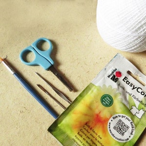 May include: A close-up of a Marabu EasyColor Batik and Fabric Dye packet, a blue pair of scissors, a blue paintbrush, and two needles. The packet is green and yellow with a white daisy and the text "EasyColor Batik und F&auml;rbefarbe" in black. The packet also has a QR code and the text "How to get your Batik Look" in a circle.