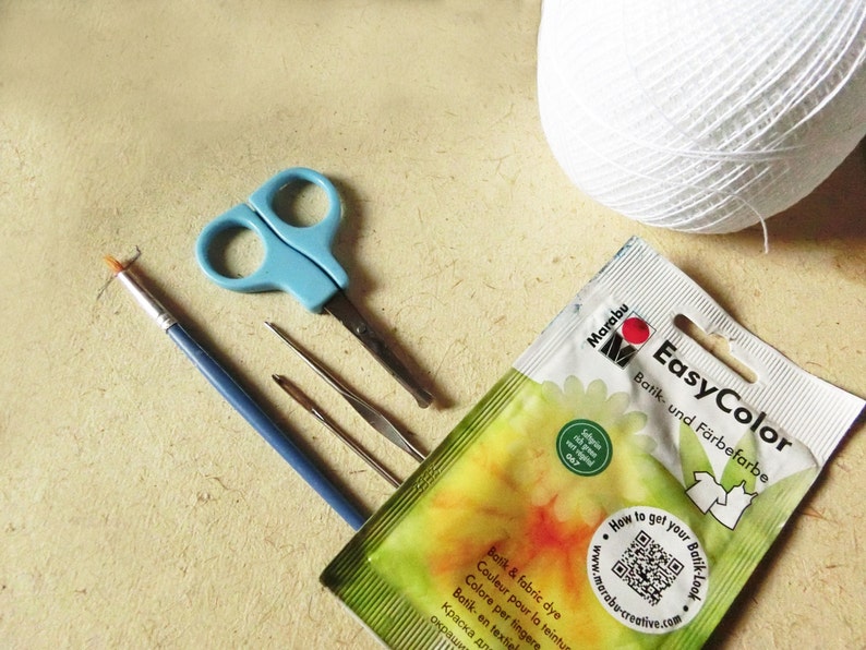 May include: A close-up of a Marabu EasyColor Batik and Fabric Dye packet, a blue pair of scissors, a blue paintbrush, and two needles. The packet is green and yellow with a white daisy and the text "EasyColor Batik und F&auml;rbefarbe" in black. The packet also has a QR code and the text "How to get your Batik Look" in a circle.
