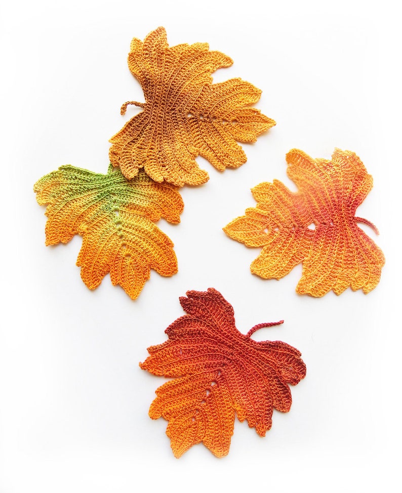 Puede incluir: Cuatro hojas de oto&ntilde;o de ganchillo en tonos de naranja, amarillo y verde. Las hojas est&aacute;n dispuestas sobre un fondo blanco.