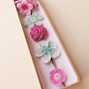 May include: A set of six crocheted flower hair clips in a brown box. The flowers are in shades of pink and green.