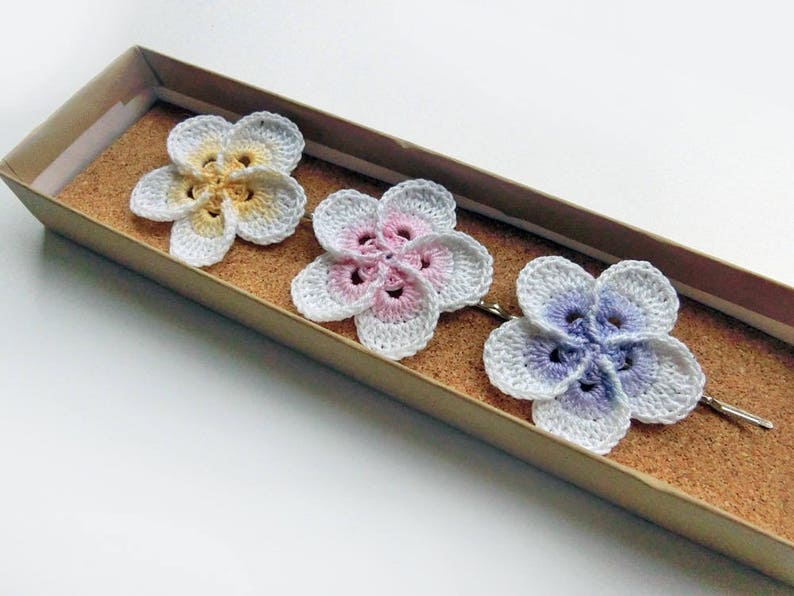 May include: Three crocheted flower hair clips in a box. The flowers are white with yellow, pink, and purple accents.
