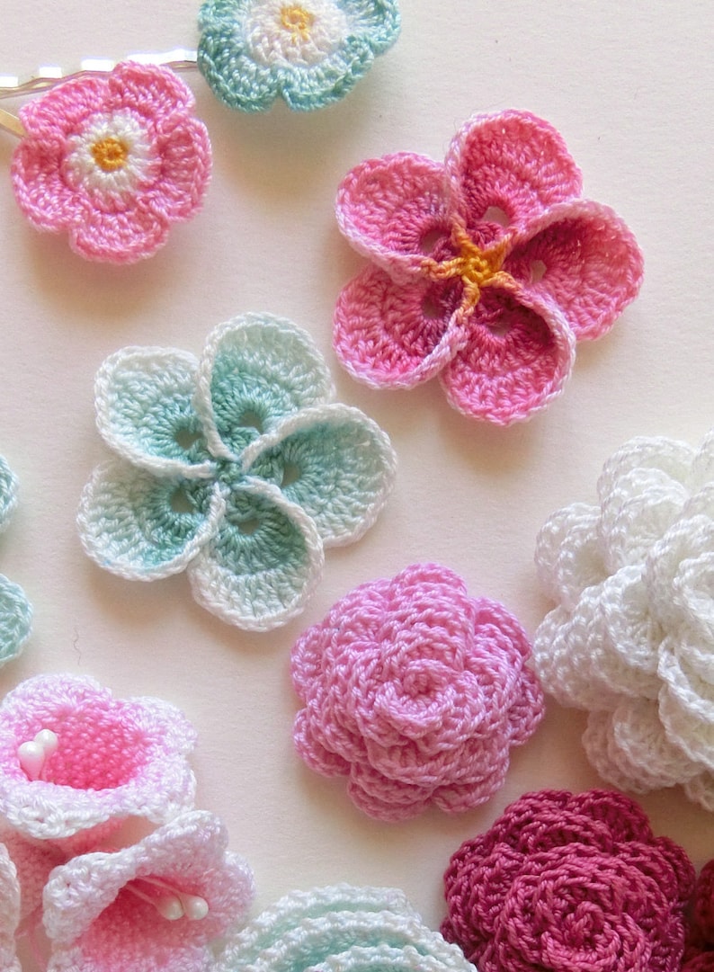May include: Assortment of crocheted flowers in various colors, including pink, white, green, and red. The flowers are made with different stitch patterns and have a delicate, handmade appearance.