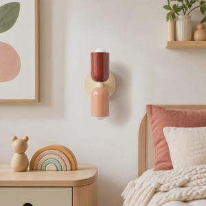 May include: A wall-mounted light fixture featuring two cylindrical shades in dark red and light peach, attached to a circular cream-coloured base. The lamp is positioned vertically on a plain wall, providing a simple and functional lighting solution.