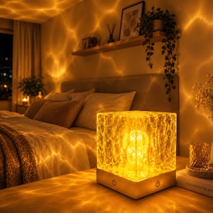 Puede incluir: Una lámpara de mesa cuadrada con una pantalla de acrílico texturizado y translúcido que emite una luz dorada cálida. La lámpara cuenta con una base sólida de color claro con dos botones de encendido en la parte frontal, creando un efecto.