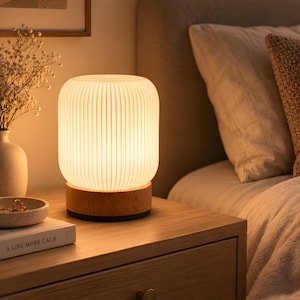 May include: A cylindrical table lamp with a vertically ribbed white shade and a light-toned wooden base. The lamp emits a warm, soft glow, illuminating the surrounding area. The design features a simple, rounded silhouette with a flat base.