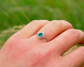 May include: A thin silver ring featuring a small, smooth, rounded piece of teal sea glass set in a simple metal bezel. The band is slender and polished, holding the vibrant, opaque teal stone securely in the centre of the ring setting.