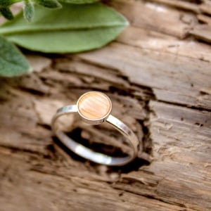 May include: A silver ring with a round, light orange stone. The ring is set on a wooden surface with green leaves and a seashell in the background. The ring's band is thin and shiny.