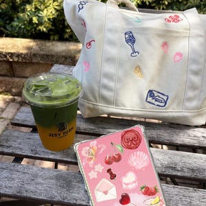 Può includere: Una borsa tote in tela color crema con icone ricamate colorate, un bicchiere di plastica trasparente contenente una bevanda ghiacciata a strati verde e arancione con il testo JELLY BEAN COFFEE e una custodia per tablet rosa con adesivi.