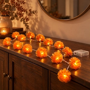 May include: A string of LED fairy lights featuring orange marigold flower covers. The lights are arranged on a wooden surface, connected by a thin wire to a clear plastic battery pack. Each flower glows with a warm, bright light.