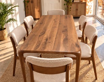 May include: A rectangular wooden dining table with rounded corners and six matching chairs. Each chair features a dark wood frame and a curved backrest upholstered in a textured, off-white fabric. The table has a smooth, natural wood grain finish.