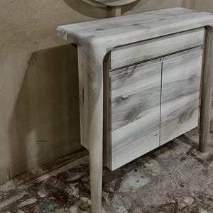 May include: A light-colored wooden console table featuring a smooth, rounded top edge that transitions into tapered legs. The unit includes one horizontal drawer positioned above two cabinet doors, all displaying a natural wood grain pattern.