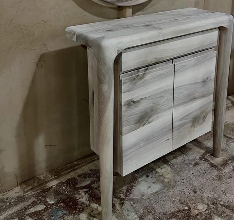 May include: A light-colored wooden console table featuring a smooth, rounded top edge that transitions into tapered legs. The unit includes one horizontal drawer positioned above two cabinet doors, all displaying a natural wood grain pattern.