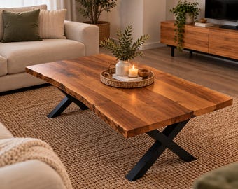 May include: A rectangular coffee table featuring a natural live edge wood slab top with a rich brown grain. The table is supported by two black metal X-shaped legs. A round woven tray sits on the surface, holding a white vase, a candle, and a small bowl.