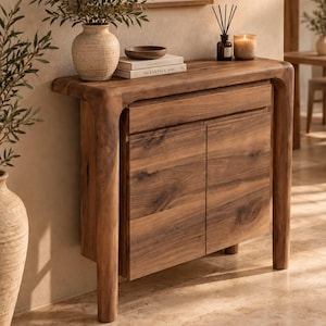 May include: A wooden console table with a smooth, rounded frame and two cabinet doors. The table features a natural wood grain finish, a single drawer above the doors, and sturdy legs. It is topped with a textured vase, books, and a candle.
