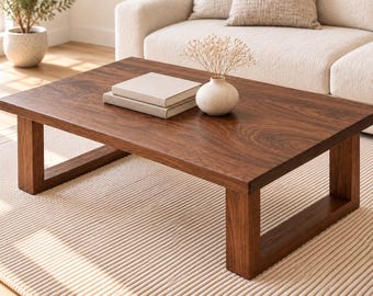 May include: A rectangular coffee table made of dark brown wood with a smooth surface and thick, U-shaped legs. Two stacked beige books and a small, round, off-white ceramic vase containing dried flowers are placed on the tabletop surface.