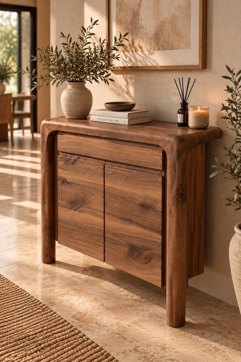 May include: A wooden console table with a smooth, rounded frame, featuring one top drawer and two cabinet doors. The natural wood grain is visible throughout the piece, which stands on four sturdy legs. The surface holds a ceramic vase, books, and a candle.