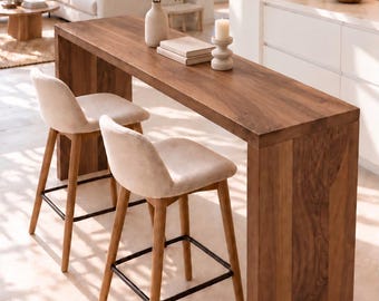 May include: A tall rectangular wooden console table with a natural grain finish, paired with two bar stools featuring light beige upholstered seats, wooden legs, and black metal footrests. The table surface holds a vase with greenery and a candle.
