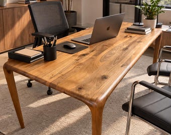 May include: A large rectangular wooden desk with a natural grain finish and rounded corners, featuring a silver laptop, a black mouse on a pad, a black pen holder with pens, and a black tray with notebooks placed on the surface for a workspace.