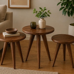 May include: A set of three round wooden side tables with tapered legs, arranged in varying heights. The tables feature a smooth, dark brown wood grain finish. A small wooden bowl sits on a book on the shortest table, while a vase and glass sit on the tallest.