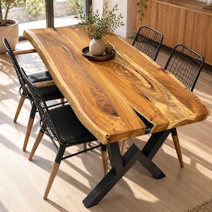 Pode incluir: Uma mesa de jantar retangular com tampo de madeira natural de borda viva com uma incrustação de resina escura, apoiada por uma base de metal preto em forma de X. Quatro cadeiras com encostos de grade preta e assentos pretos rodeiam a mesa.