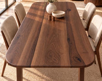 May include: A rectangular walnut wood dining table with rounded corners and a sturdy base, accompanied by four chairs featuring light beige upholstered seats and wooden frames, arranged in a bright, open living space.