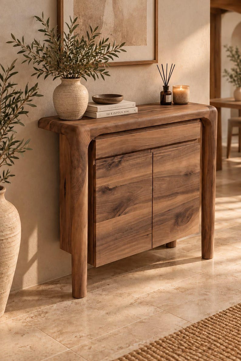 May include: A wooden console table with a smooth, rounded frame and two cabinet doors. The table features a natural wood grain finish, a single drawer above the doors, and sturdy legs. It is topped with a textured vase, books, and a candle.