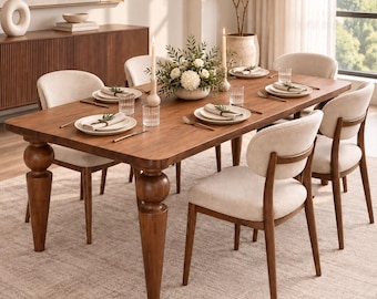 May include: A rectangular wooden dining table with four thick, rounded legs and six upholstered chairs with curved backrests. The table is set with white plates, clear glasses, gold-toned cutlery, linen napkins, and a central floral centerpiece.