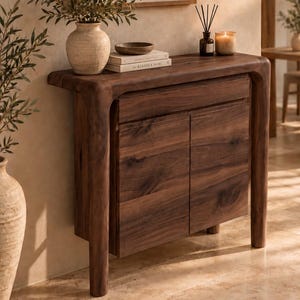 May include: A dark brown wooden console table featuring a smooth top, a single drawer, and two cabinet doors. The table has rounded edges and thick, curved legs. On top sits a textured beige vase with greenery and a book titled The Kinfolk Home.