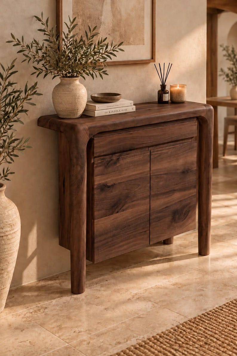 May include: A dark brown wooden console table featuring a smooth top, a single drawer, and two cabinet doors. The table has rounded edges and thick, curved legs. On top sits a textured beige vase with greenery and a book titled The Kinfolk Home.