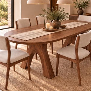 Peut inclure: Une table de salle à manger ovale en bois avec une base arquée épaisse et six chaises dotées d'assises et de dossiers rembourrés beige clair. Un chemin de table rayé est posé sur la surface, avec un vase sombre, de la verdure et deux bougies.