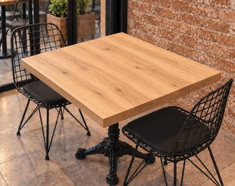 May include: A square wooden table with a light brown grain finish and a black metal pedestal base, accompanied by two black wire-frame chairs featuring black seat cushions. The table surface is smooth and flat, supported by a decorative base.