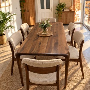 Puede incluir: Una mesa de comedor rectangular de madera oscura con esquinas redondeadas y seis sillas con marcos de madera oscura y tapicería de tela texturizada de color beige claro. Un florero de cristal con flores blancas sobre un mantel individual trenzado.