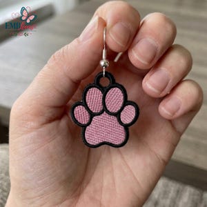 May include: A dangling earring featuring a pink embroidered paw print design with a black stitched border. The paw shape is attached to a silver-toned metal fishhook ear wire, creating a lightweight accessory with a textured fabric finish.