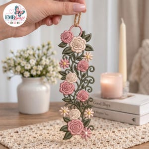 Può includere: Un ornamento da appendere ricamato in verticale con un motivo floreale di rose rosa e crema, piccole margherite e foglie verde scuro a spirale. Il pezzo è realizzato con un intricato lavoro di filo e include un piccolo occhiello per appenderlo.