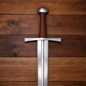 May include: A steel sword featuring a double-edged blade with a central fuller, a straight crossguard, and a brown leather-wrapped grip topped with a rounded metal pommel. The weapon displays a polished metallic finish and a symmetrical design.