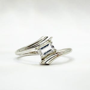 May include: A silver-toned ring featuring a single rectangular clear gemstone set between two bypass-style bands that curve around the stone. The metal has a polished finish and the design creates a split shank appearance on both sides.