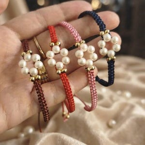 May include: Four braided cord bracelets in brown, red, pink, and navy blue. Each bracelet features a central flower motif composed of five white round pearls accented with small gold-toned metal beads at the center and edges of the petals.