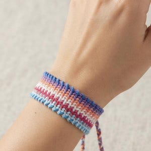 May include: A woven friendship bracelet featuring a horizontal striped pattern in shades of purple, pink, white, orange, and light blue. The bracelet has a wide, flat band design with two braided ties extending from the bottom, each ending in a small knot.