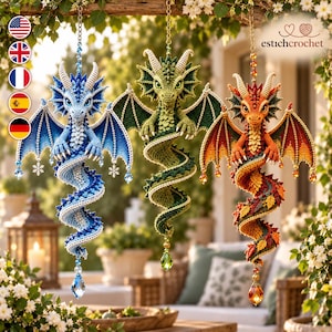 May include: Three hanging crochet dragon ornaments in blue, green, and orange. Each dragon features detailed scales, wings, and a coiled tail, suspended by a chain with a dangling crystal pendant. The blue dragon has snowflake accents.