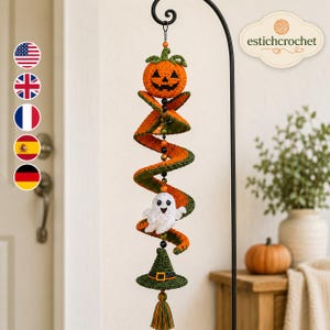 Op de afbeelding: Een hangende gehaakte Halloween-decoratie met een spiraalvormige structuur van oranje en groen garen. Het bevat een pompoen aan de bovenkant, een klein wit spookje in het midden en een groene heksenhoed aan de onderkant, versierd met kralen.