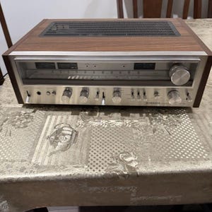 Pode incluir: Um recetor estéreo Pioneer vintage com painel frontal em metal prateado, vários botões de controlo, interruptores de alavanca e um mostrador de rádio coberto por vidro. A unidade está inserida numa caixa de madeira escura com grelha preta.